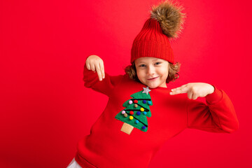 cool teenager in a red Christmas sweater fooling around against the background of a red wall, a warm hat and a sweater with a Christmas tree