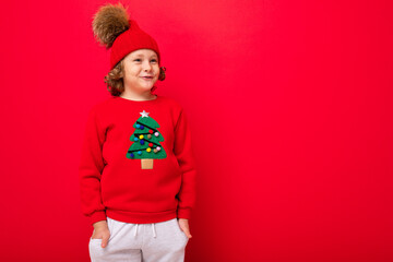 cool teenager in a red Christmas sweater fooling around against the background of a red wall, a warm hat and a sweater with a Christmas tree