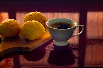 Xícara branca de café e pães de queijo sobre superfície reflexiva.
