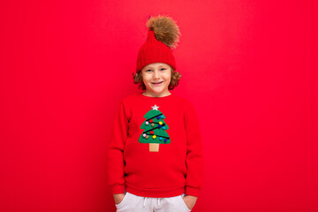 cute blond boy in warm hat and christmas sweater on red background with smile on his face