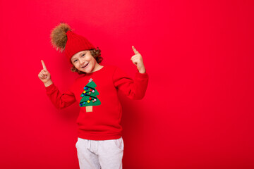 cool teenager in a red Christmas sweater fooling around against the background of a red wall, a warm hat and a sweater with a Christmas tree