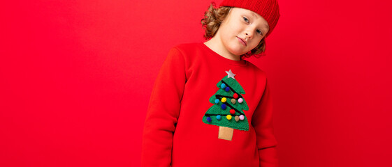 cool teenager in a red Christmas sweater fooling around against the background of a red wall, a warm hat and a sweater with a Christmas tree