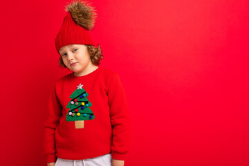 cute blond boy in warm hat and christmas sweater on red background with smile on his face