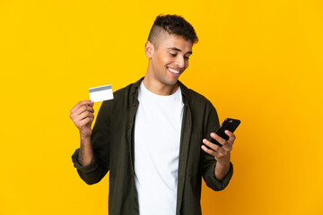 Young brazilian man isolated on yellow background buying with the mobile with a credit card