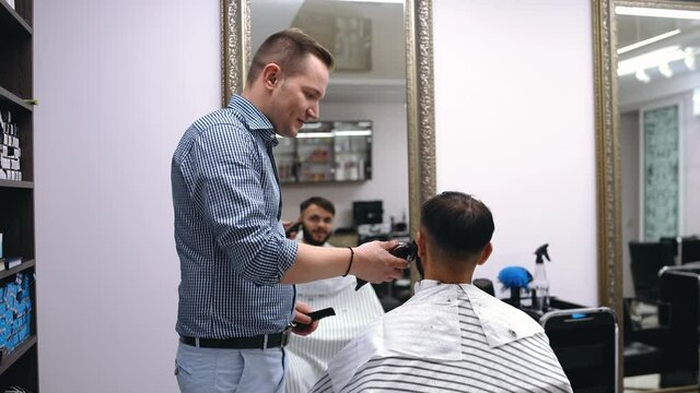 Young man in barber shop sits in an armchair. Barber cuts his hair with a trimer.
