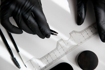 the master in black vinyl gloves holds a pair of tweezers for making eyebrows on the background of a ruler, brushes.