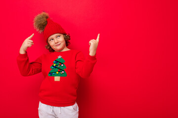 cool teenager in a red Christmas sweater fooling around against the background of a red wall, a warm hat and a sweater with a Christmas tree