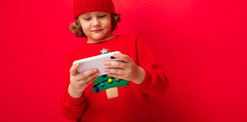 a young man with a smartphone in his hands against the background of a red wall, in a knitted hat and a sweater with a Christmas tree.