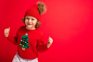 cool blond kid in warm hat and sweater with christmas tree on red background fooling around, christmas concept