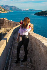 Attractive brunette traveler posing on the old city walls of the town Dubrovnik, tourist exploring the ancient city on a warm autumn day. Wearing a white shirt and black leather pants