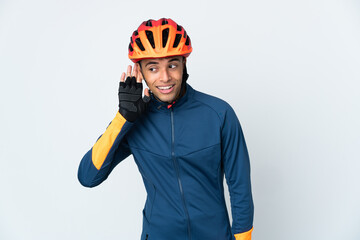 Young cyclist Brazilian man isolated on white background listening to something by putting hand on the ear