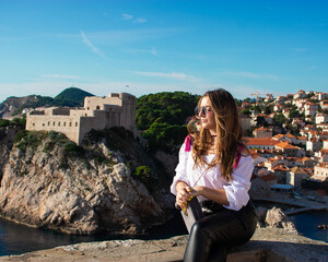 Fototapeta na wymiar Attractive brunette traveler posing on the old city walls of the town Dubrovnik, tourist exploring the ancient city on a warm autumn day. Wearing a white shirt and black leather pants
