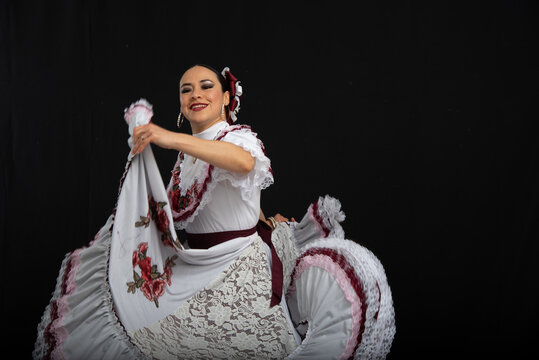 Bailarina mujer latina vistiendo traje de Aguascalientes México, fondo negro, con vestido blanco con cintas color vino, moviendo su falda y sonriendo