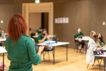 Rear view of an unrecognizable woman with red hair holding a presentation indoors, people inside sitting with space in between them, following covid guidelines while wearing facemasks