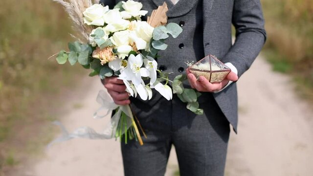 Portrait of young stylish groom holding beautiful white wedding bouquet and rings on sunny background outside. Elegant male person smiling looking at camera. Marriage party decoration details. Youth.