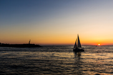Fototapeta premium Silhouette of a sailboat on the sea next to a dock with a sunset on the horizon.