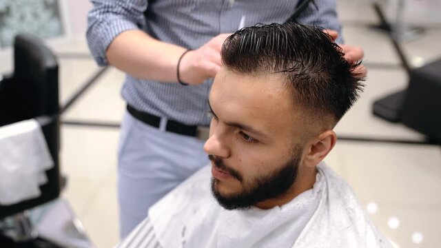 Unparalleled barber with a beard and a tattoo is cutting the hair of his client in the barbershop. He is using a cutting comb and a hair clipper.
