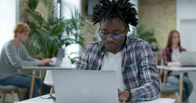 Focused young African American man surfing Internet on laptop with blurred colleagues working online at the background. Handsome millennial guy analyzing strategy in office. Cinema 4k ProRes HQ.
