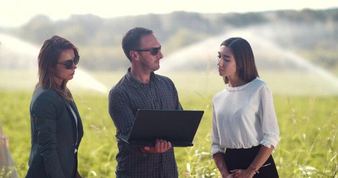 Business people shaking hands after successful presentation on laptop in a green field with agriculture irrigation system. Water sprinklers in the background