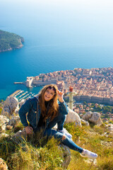 Naklejka premium Vertical shot of a brunette sitting on the stones on the Srd mountain above the city of Dubrovnik, posing for a photo and laughing as she holds her sunglasses