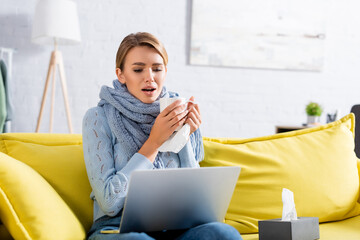 Diseased freelancer with cup and napkin having video chat on laptop on couch