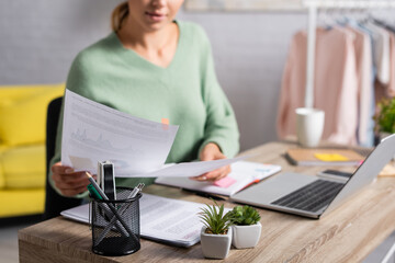 Plants and stationery on table near freelancer holding papers and laptop on blurred background