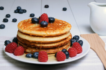 Plato de tortitas americanas con sirope de arce, arándanos y frambuesas sobre mesa de madera blanca y con jarra de porcelana y cubierto de madera.