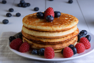 Plato de tortitas con sirope de arce, arándanos y frambuesas sobre mesa de madera blanca con frutos esparcidos y desenfocados.