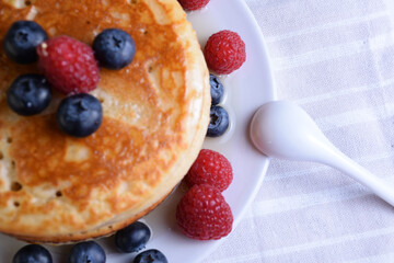 Plato de tortitas americanas con sirope de arce, arándanos y frambuesas semi-desenfocado sobre tela clara y cuchara de porcelana, perspectiva cenita y parcial.