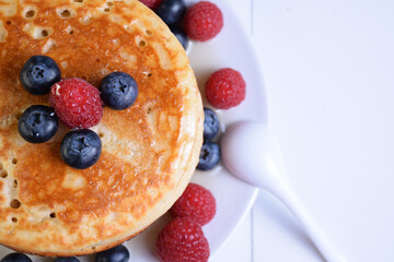 Plato de tortitas americanas con sirope de arce, arándanos y frambuesas semi-desenfocado sobre mesa de madera blanca y cuchara de porcelana, perspectiva cenita y parcial.