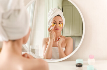 Obraz na płótnie Canvas Young female standing near mirror and applying cosmetic collagen yellow under eye patches during morning skincare procedure in bathroom.