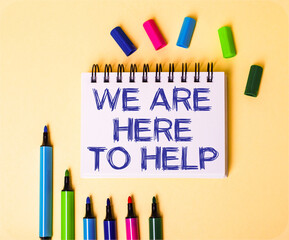 WE ARE HERE TO HELP written in a white notebook on a beige background near multi-colored markers
