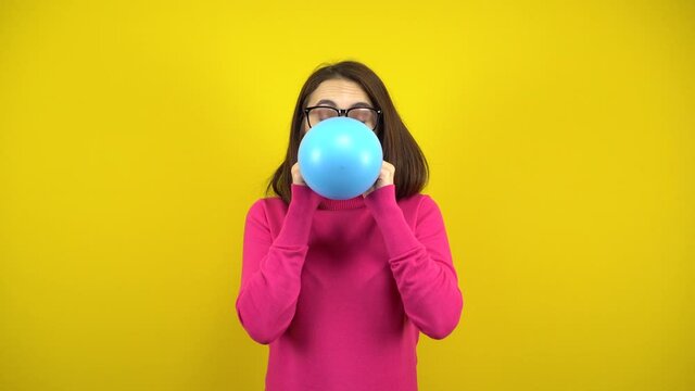 A young woman inflates a blue balloon with her mouth on a yellow background. Girl in a pink turtleneck and glasses.