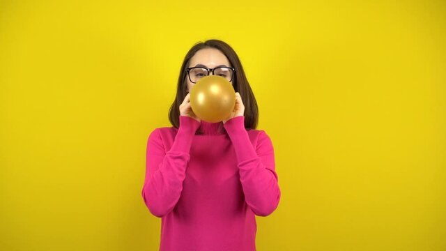 A young woman inflates a gold balloon with her mouth on a yellow background. Girl in a pink turtleneck and glasses.