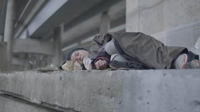 Female refugee covered with dirty coat sleeping under the bridge, unemployment