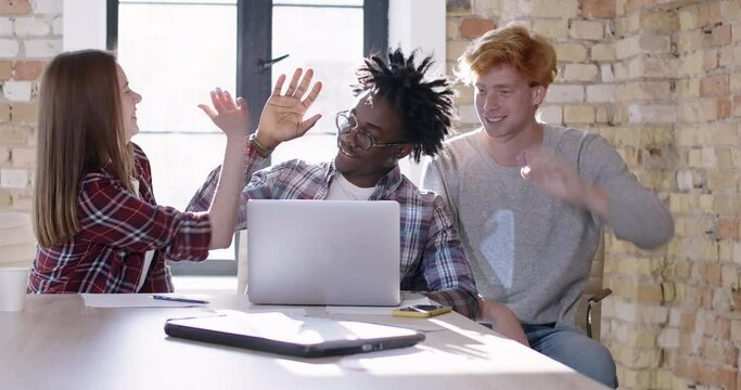 Three happy coworkers showing victory gesture and giving high-five in sunny office. Portrait of cheerful millennial multiethnic young colleagues rejoicing successful startup. Cinema 4k ProRes HQ.