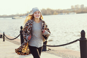 blond teenager girl in knitted hat and trench jogging on the city river background