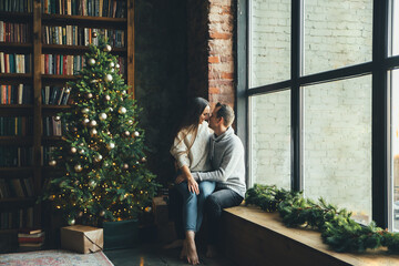 Couple in love near festive Christmas tree. A beautiful young wife and her husband sit on the floor in tenderly embrace. New Year eve together