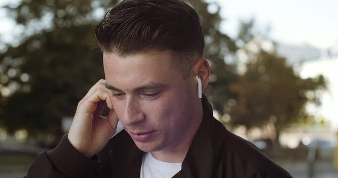 Portrait of young Caucasian student cheerful man with wireless headphone in ear having phone call and actively speaking smiling standing outdoor. Stylish guy talk using smartphone for communication