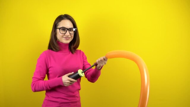 A young woman inflates a long orange balloon with a pump on a yellow background. Girl in a pink turtleneck and glasses.