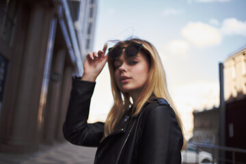 Fototapeta premium Fashionable woman in a leather jacket and sunglasses near the building on the street