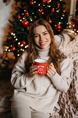 Young woman smiling, relaxing at home, enjoying Christmas. Beautiful girl drinking tea at home, sitting near the Christmas tree