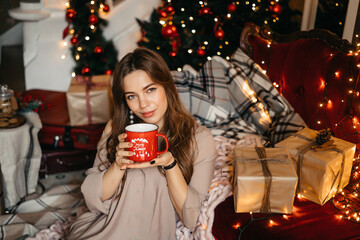 Young woman smiling, relaxing at home, enjoying Christmas. Beautiful girl drinking tea at home, sitting near the Christmas tree