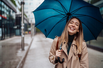 A beautiful smiling young woman walking through the city with an umbrella. - obrazy, fototapety, plakaty