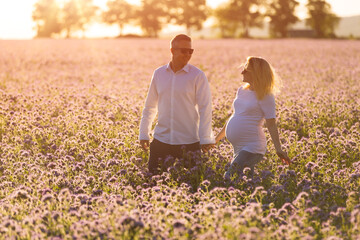 Beautiful pregnant mother and her husband walk together hand in hand outdoor, in the middle of a flowery field at sunset time.