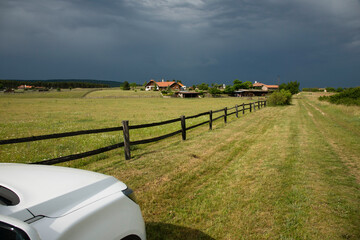Rural view, stormy clouds and green field with white car and wooden corral like fence.