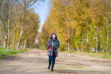 new normal Autumn walk outdoors - young happy and pretty Asian Japanese woman in face mask walking cheerful at beautiful city park in vibrant yellow and orange tree leaves