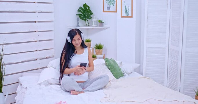 Pregnant woman sits on the bed with headphones and listens to music. Woman looks at the phone and strokes her tummy with her hand, happy pregnancy. 4k, ProRes