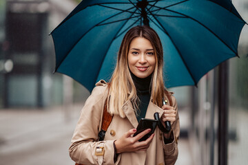 Fashionable smiling young woman under an umbrella holding a mobile phone.