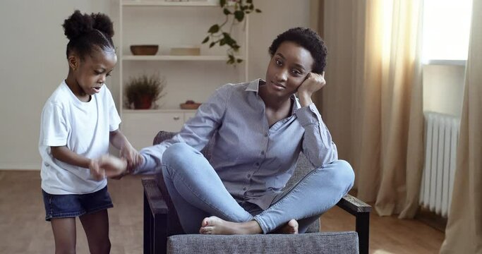 Young tired single african mother suffers from headache sitting on couch feeling stress while her daughter running around her shouting touching mom, black female babysitter exhausted by noisy kid girl